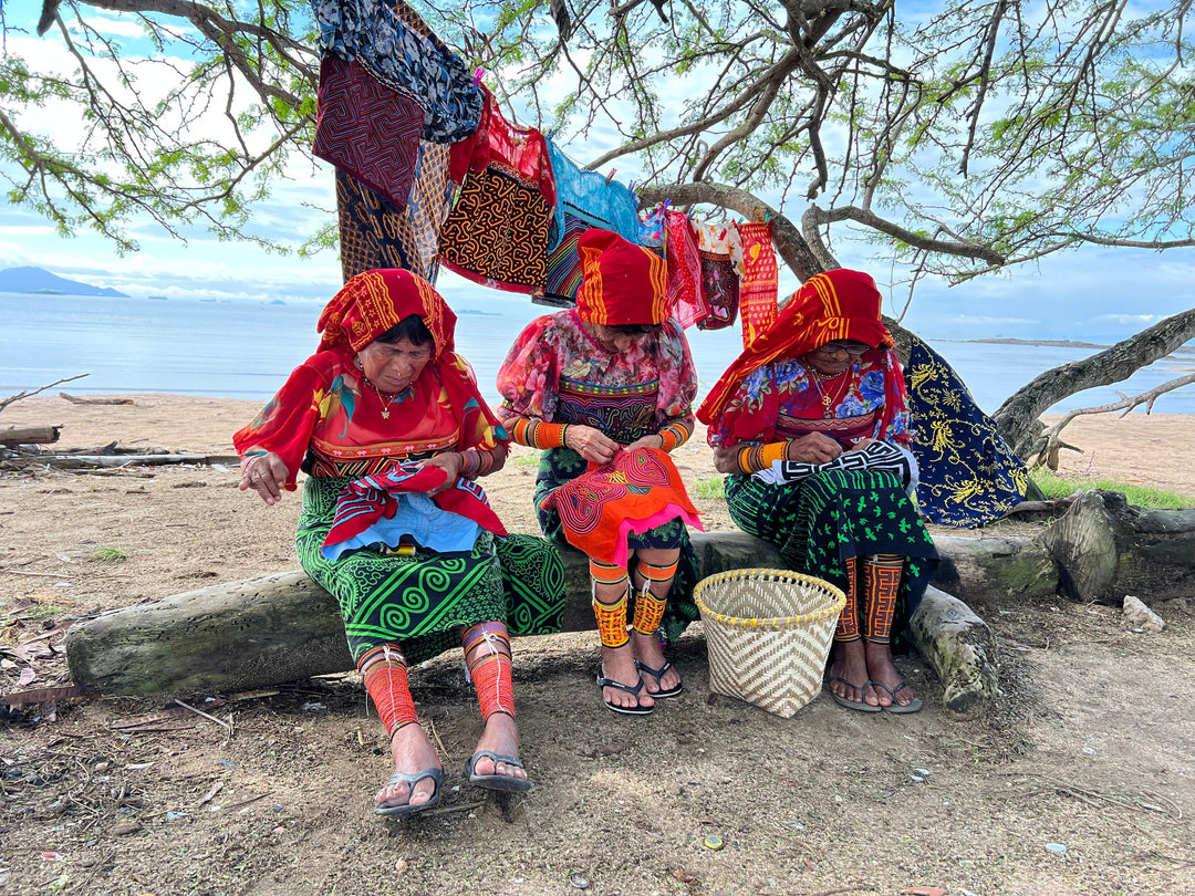 Three Guna women sit on a log near a beach, dressed in traditional vibrant clothing and headscarves, working on handmade molas. Colorful molas hang on a tree branch above them, with the ocean and sky in the background.