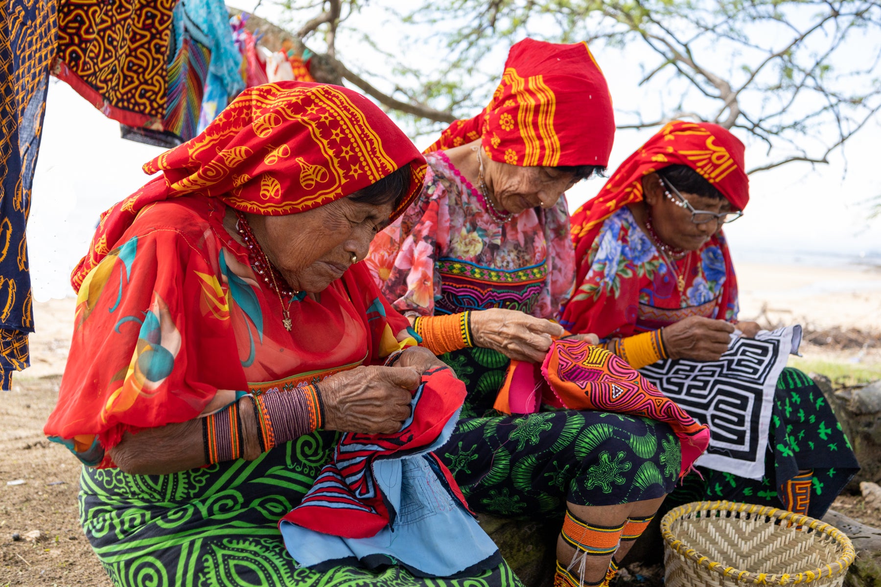 Three Kuna women skillfully sewing a vibrant mola, a traditional textile art, for sale on TraderBrock.com, where you can find unique handcrafted items.